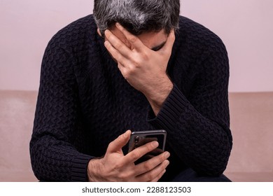 Middle-aged man receiving sad news on the phone. Concerned man reading news online looking at phone screen, Caucasian sitting on sofa at home, serious and sad - Powered by Shutterstock