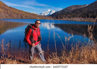 A middle-aged man in a red jacket and glasses with a backpack for tourism on the background of the lake which reflects the snow-capped mountains. - Powered by Shutterstock