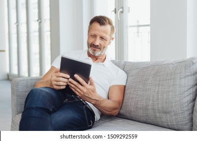 Middle-aged Man Relaxing On A Sofa With A Tablet Computer Smiling Quietly As He Reads Something On The Screen In A Low Angle View With Copy Space