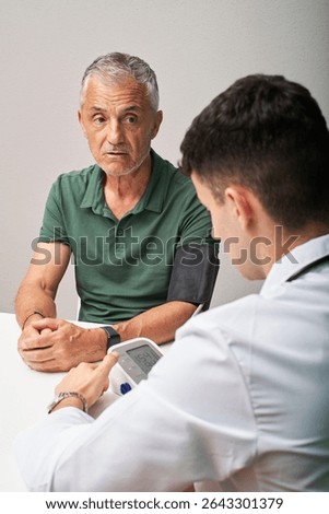 A middle-aged man sits with a concerned expression while a young doctor measures his blood pressure during a routine health check-up in a modern clinic.
