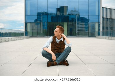 Middle-aged man sits cross-legged on rooftop in urban area, looking thoughtfully to side. Tall buildings surround him. Bright daylight shines down, revealing clear sky.