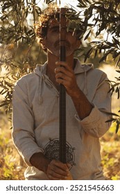 A middle-aged man sits on the ground in an olive field, holding his baglama close, his eyes closed in reflection. The sun filters through the olive branches above