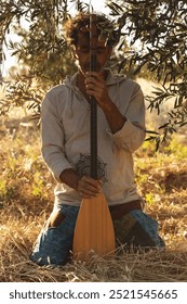 A middle-aged man sits on the ground in an olive field, holding his baglama close, his eyes closed in reflection. The sun filters through the olive branches above