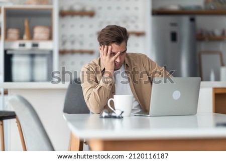 Middle-aged man sitting in front of laptop in dining room and feels stressed after realized how much work he has and that he will not be able to go on vacation.