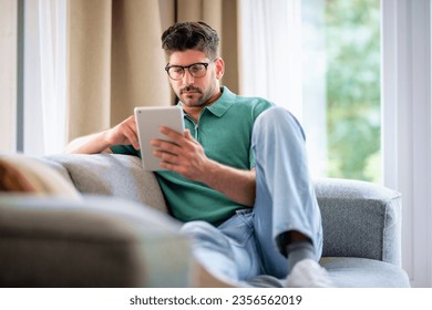 Middle-aged man sitting at home on the sofa holding a digital tablet in his hand. Confident male wearing glasses and casual clothes. He is having video call or browsing on the internet. Full length - Powered by Shutterstock