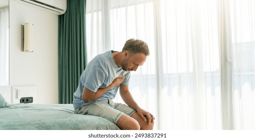 Middle-aged man sitting on bed, clutching chest in pain, heart attack or respiratory issue - Powered by Shutterstock