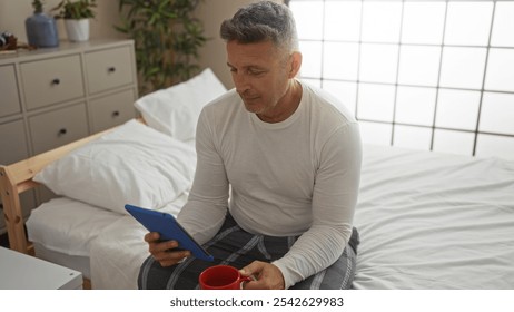 Middle-aged man sitting on bed in bedroom holding tablet and red mug while wearing casual clothes - Powered by Shutterstock