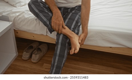 Middle-aged man sitting on bed, holding his foot, wearing pajamas, in a bedroom with wooden floor and slippers nearby, indicating morning routine or foot discomfort in a home setting - Powered by Shutterstock