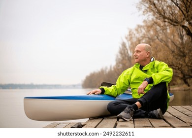 A middle-aged man sitting on the jetty and ready to go kayaking. Active sporty man wearing dry suit and looking away.  - Powered by Shutterstock