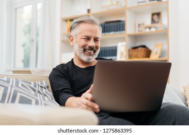 Middle-aged Man Sitting On The Sofa And Smiling Happy While Watching A Funny Video On The Internet On His Laptop At Home