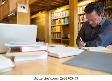 Middle-aged man sitting at study table in library writing in notebook beside laptop, copy space. Intellectual, focus, concentration, education, workspace, academic, professional