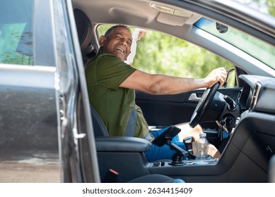 A middle-aged man smiles while sitting in the drivers seat of a car, enjoying a travel adventure. He appears relaxed and content, taking in the warm weather and scenic surroundings.