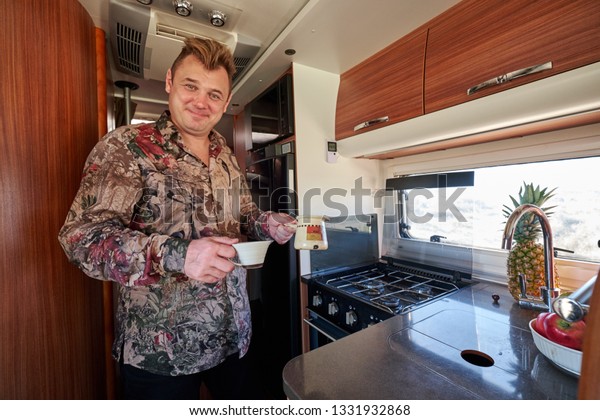 Middle-aged man smiling looking at camera standing\
inside of recreational vehicle motor home kitchen corner, holding\
cup and coffee in cezve or jezve starting day drinking hot beverage\
concept image