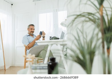 Middle-aged man smiling while using his mobile phone at his home office.