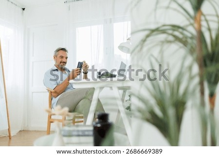 Middle-aged man smiling while using his mobile phone at his home office.