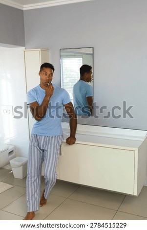 Middle-aged man standing and brushing teeth with toothbrush in bathroom, leaning on floating vanity. Mirror, toilet, cabinet, tiles, minimalist, modern, sunlit