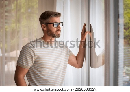 A middle-aged man standing at the window, deep in thought. Confient male with gray hair and glassses looking out the window. 