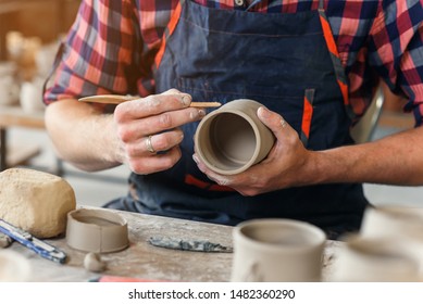 A middle-aged man in a uniform work by wooden stack with a clay shape of cup in a large creative pottery manufacture. Artwork and handmade concept. - Powered by Shutterstock