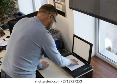 Middle-aged man using a flatbed scanner in an office. Employee scans a document on the desk near a window, workplace technology, digitalization, paperwork, copying, business routine, office job scene.
