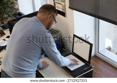 Middle-aged man using a flatbed scanner in an office. Employee scans a document on the desk near a window, workplace technology, digitalization, paperwork, copying, business routine, office job scene.
