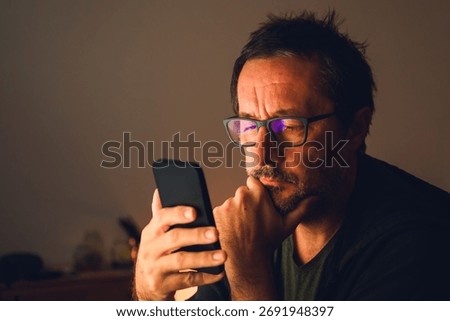 Middle-aged man using smartphone in dimly lit room, focused on screen with reflection of blue light in glasses. Selective focus.