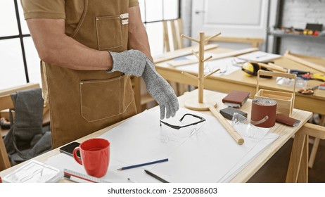 A middle-aged man wearing gloves stands by a workbench in a well-equipped carpentry workshop. - Powered by Shutterstock