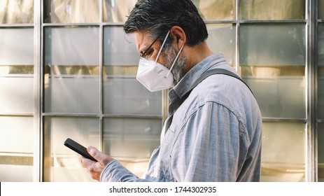 A middle-aged man wearing a KN95 (or FFP2) face mask consults his mobile as he walks past aehead the window of a closed business.  - Powered by Shutterstock