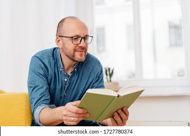 Middle-aged Man Wearing Reading Glasses Sitting In A Chair In His High Key Living Room Reading A Book With A Quiet Smile Of Pleasure