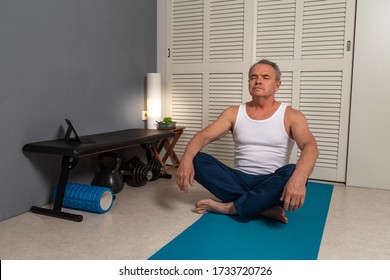 Middle-aged Man Wearing A White Tank Top And Blue Sweat Pants Practicing Yoga On The Mat At Home