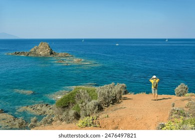 A middle-aged man wearing a yellow T-shirt and carrying a backpack takes pictures of a beautiful landscape while hiking along the Italian coast. Falcon Bay, Piombino, Tuscany. - Powered by Shutterstock