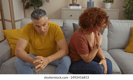 Middle-aged man and woman sit on couch in living room, both looking upset and distant, depicting a couple's serious relationship issue at home. - Powered by Shutterstock