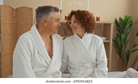 Middle-aged man and woman wearing white robes, smiling at each other in a serene spa room with candles and plants - Powered by Shutterstock