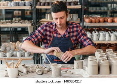 A middle-aged man in a work uniform wipes with a damp sponge a clay shape of cup in a large creative pottery workshop. Art workspace concept. - Powered by Shutterstock