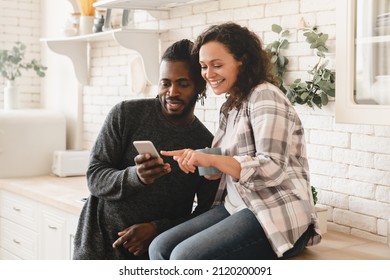 Middle-aged Mature Happy African Couple Using Smart Phone Cellphone For Surfing Social Media, Sharing Photos, Checking E-mail, Using Mobile Application Online Relaxing At Home Kitchen