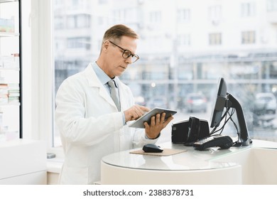 Middle-aged mature male caucasian chemist druggist pharmacist using digital tablet standing at cash point desk in pharmacy drugstore looking for medicines online - Powered by Shutterstock