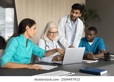 Middle-aged and millennial internists professionals using laptop, review diagnosis, check-up results of patient, gathered for decision making in modern meeting room. Teamwork, medicine and modern tech