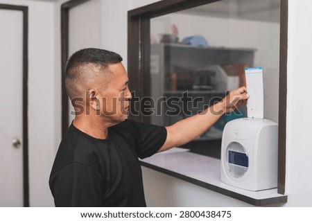 A middle-aged office worker inserts attendance card into a time recorder machine to clock in for work, tracking employee hours and workplace attendance.