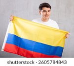Middle-aged positive man in casual clothes holds unfurled flag of Colombia in hands raised above head against gray wall, studio shot. Support, national identification