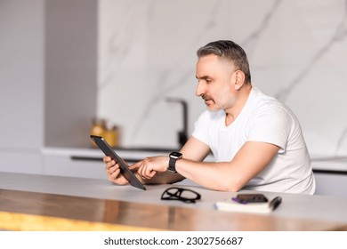 Middle-aged professional man looking at camera and checking messages on digital tablet while having breakfast on kitchen counter at home - Powered by Shutterstock