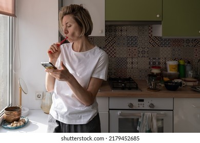 Middle-aged Scandinavian woman brushing teeth in morning and using smartphone, mature lady holding toothbrush and scrolling social media on mobile phone while standing by window in kitchen - Powered by Shutterstock