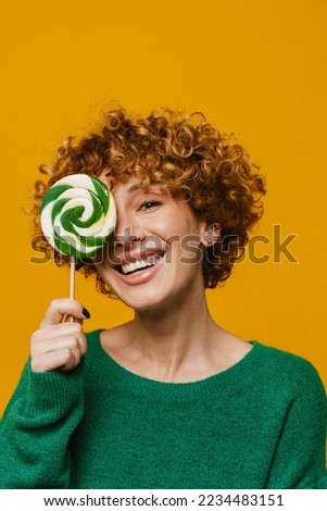 Middle-aged smiling ginger woman covering her eye with lollipop isolated over yellow studio background