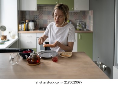 Middle-aged unkempt woman spreads jam on toast having breakfast alone. Mature blonde female eats healthy food in morning thinking about own lifestyle. Lady prepares humble breakfast in late morning - Powered by Shutterstock