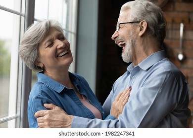 Middle-aged untroubled couple happy homeowners hugging laughing while standing in kitchen at home. Medical insurance cover, dental clinic services for older citizen, carefree midlife and love concept
