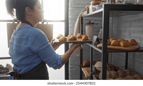 Middle-aged woman arranging freshly-baked croissants on a shelf in a cozy bakery, showcasing a warm indoor setting filled with a variety of pastries and bread. - Powered by Shutterstock