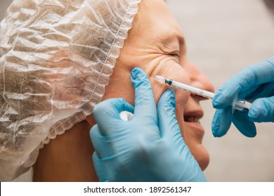 Middle-aged woman with crow's feet wrinkles around her eyes undergoing rejuvenation procedure using hyaluronic acid filler injections. Cosmetologist injects botulinum toxin for smooth face skin.