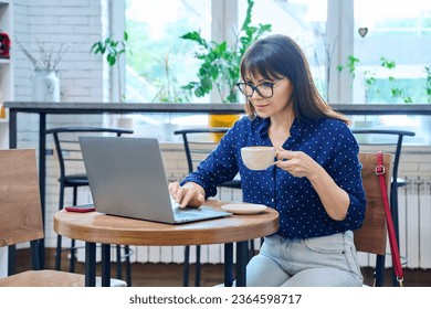 Middle-aged woman drinking coffee looking at laptop screen, sitting in coffee shop - Shutterstock ID 2364598717