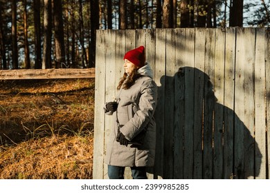 A middle-aged woman exudes autumn elegance as she strikes a pose by an old, shabby fence on a sunny day, showcasing her stylish fall attire in a charming rustic setting. - Powered by Shutterstock