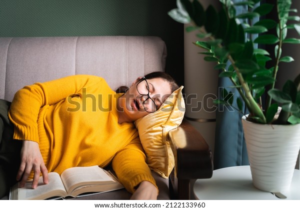 Middle-aged woman in\
glasses fell asleep while reading book at home, female sleeping\
with open mouth on sofa near coffee table with houseplant in living\
room. Sleep\
deprivation