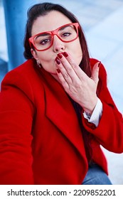 Middle-aged Woman With Glasses Using A Phone To Take A Selfie. She Is Dressed In A White Blouse, Blue Jeans And Red Coat. Defocused Background Of An Office Building. Business Executive Woman Concept.