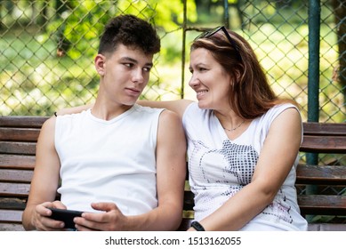 A Middle-aged Woman Is Good Spending Time With Her Son In A Park, They Sit And Talk. Mom Looks At Her Teenage Son With Love. Family Concept